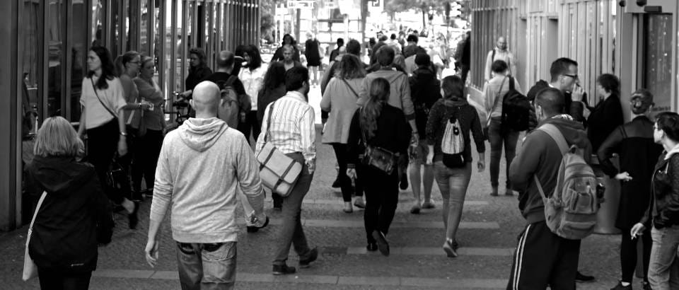 A black and white street scene showing a crowd of people walking along a city sidewalk between two buildings.